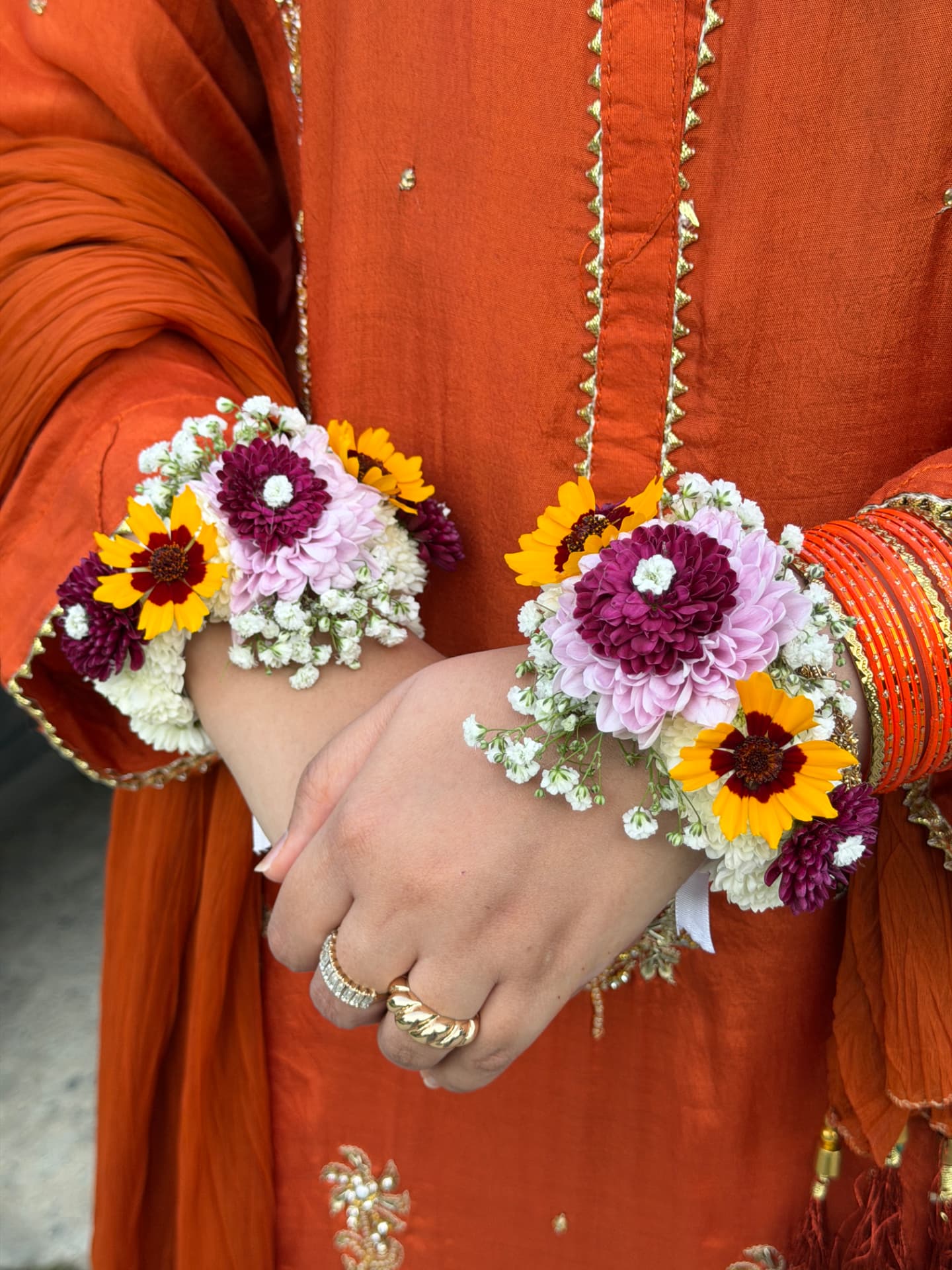 Two wrists wearing plum, lilac and yellow daisy floral bracelets against a rust orange mehndi outfit