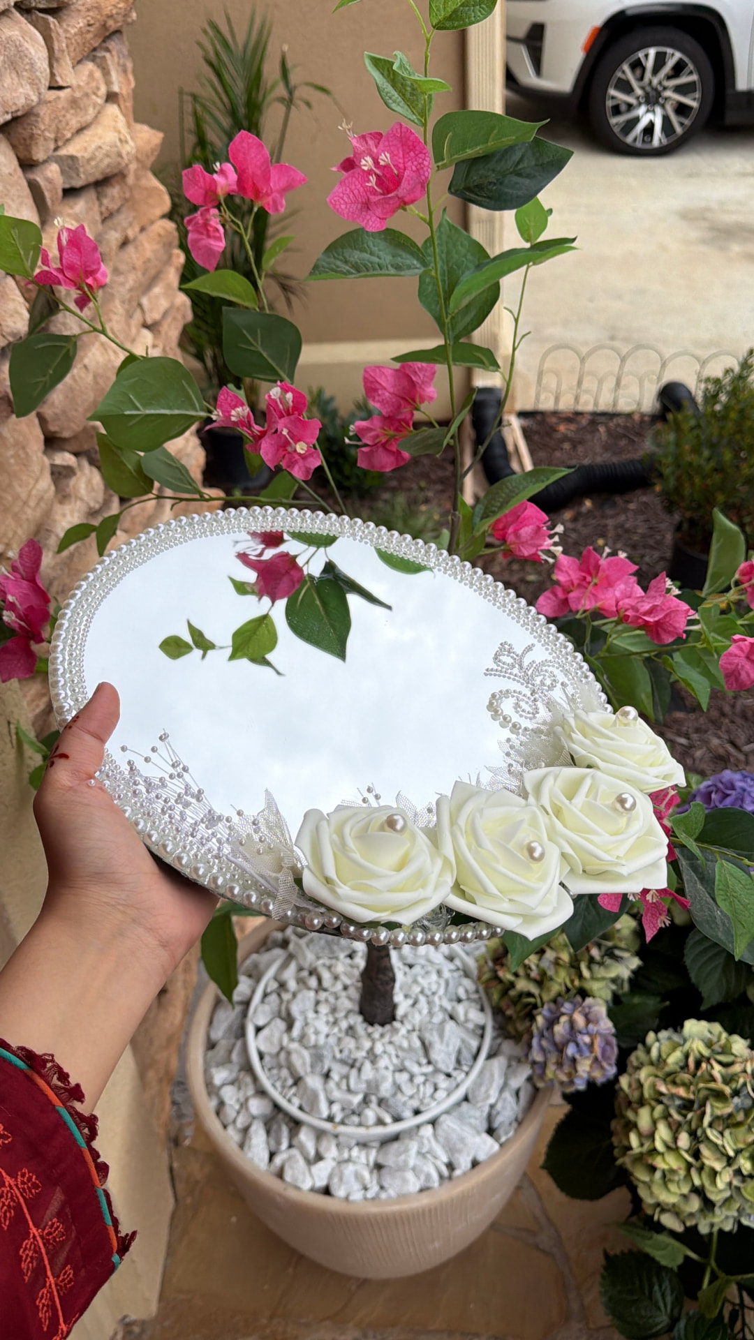 Hand holding an oval bridal mirror trimmed with pearls and ivory roses in front of pink bougainvillea