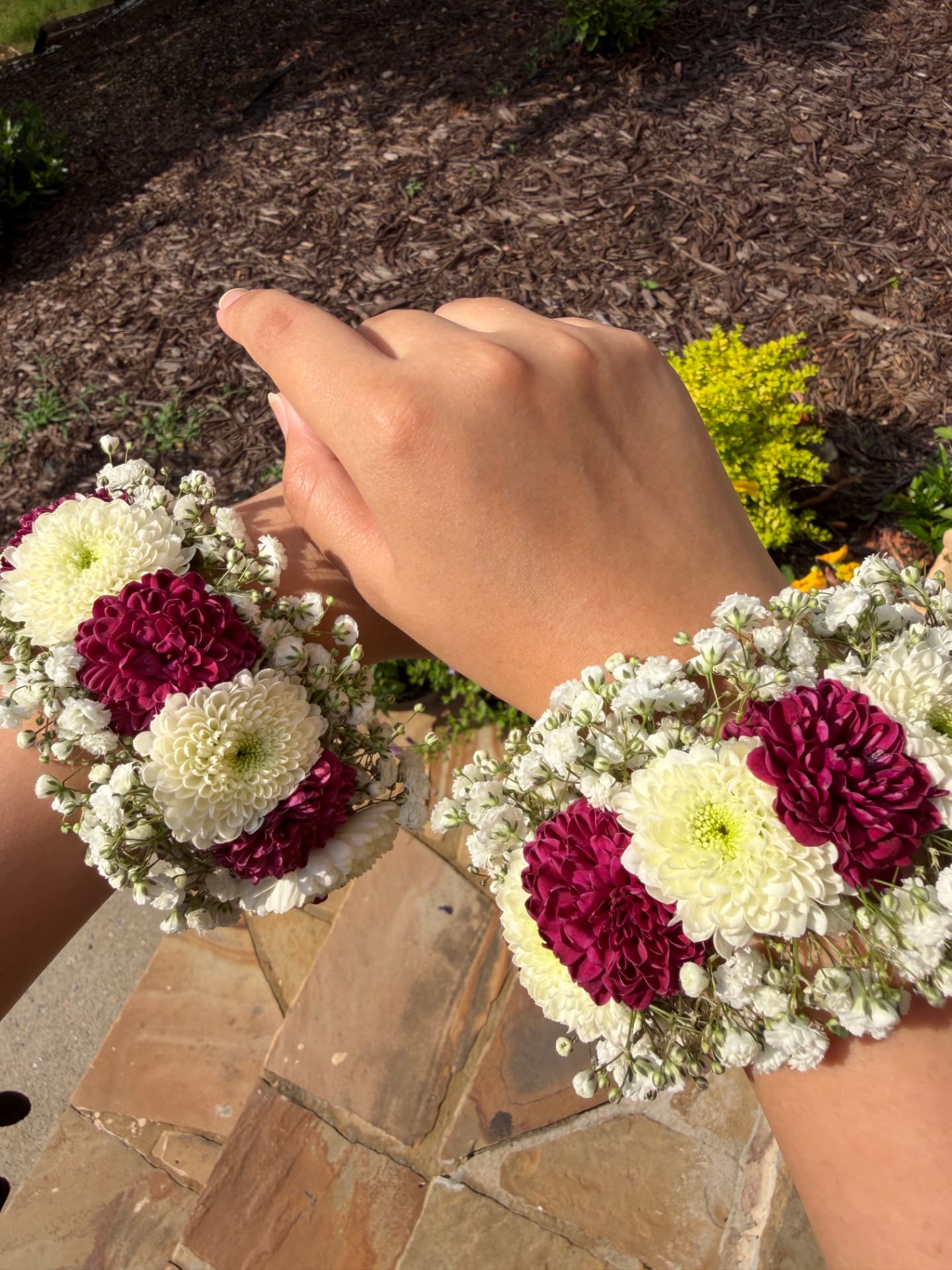 Two matching floral bracelets of burgundy and ivory chrysanthemums with baby's breath worn on both wrists outdoors