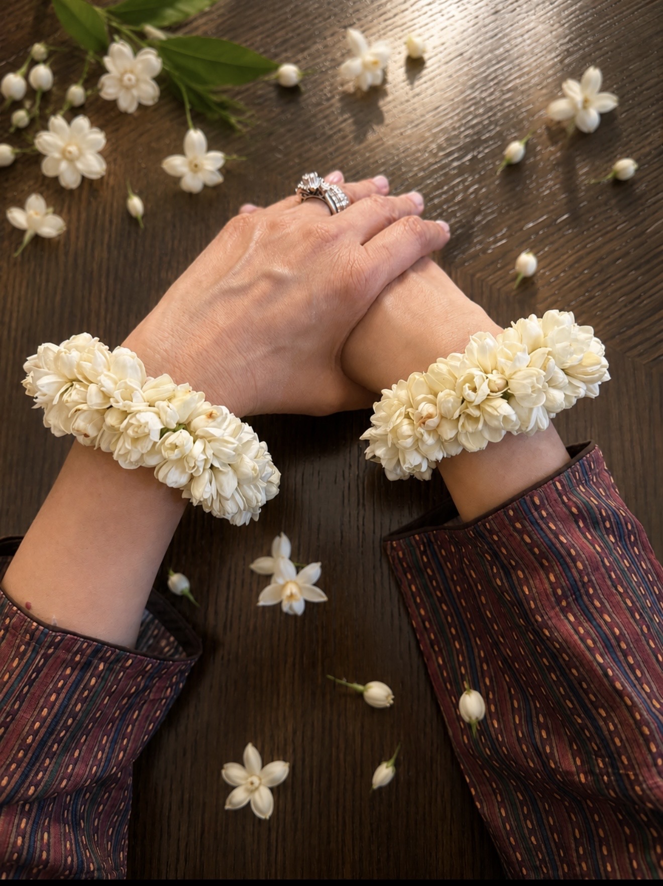 Two pairs of hands resting on dark wood wearing full fresh jasmine flower gajras, with loose jasmine blooms scattered around